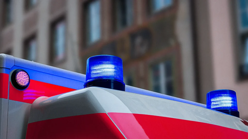 Ein Teil eines Rettungswagens mit zwei blauen Warnleuchten auf dem Dach vor unscharfem städtischem Hintergrund.