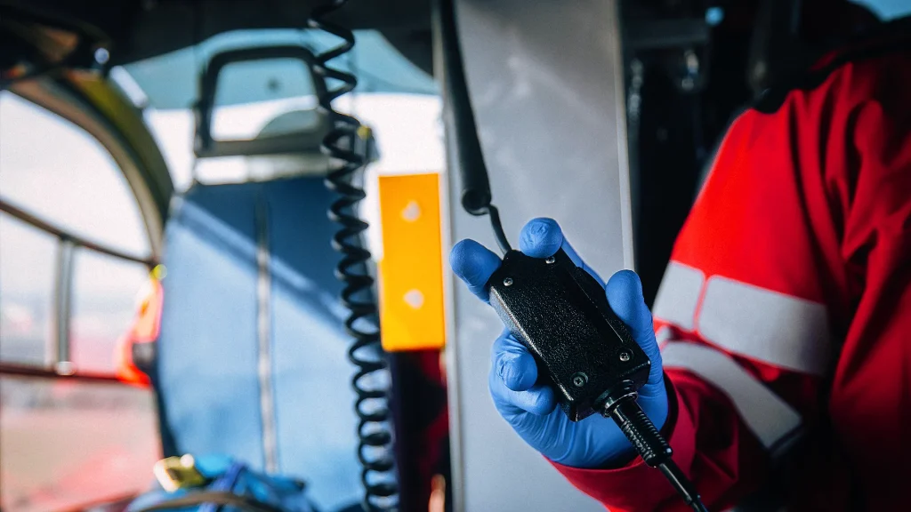 Eine Person in roter Schutzkleidung und blauen Handschuhen hält ein Funkgerät in der Hand in einem Hubschrauber mit technischen Geräten.
