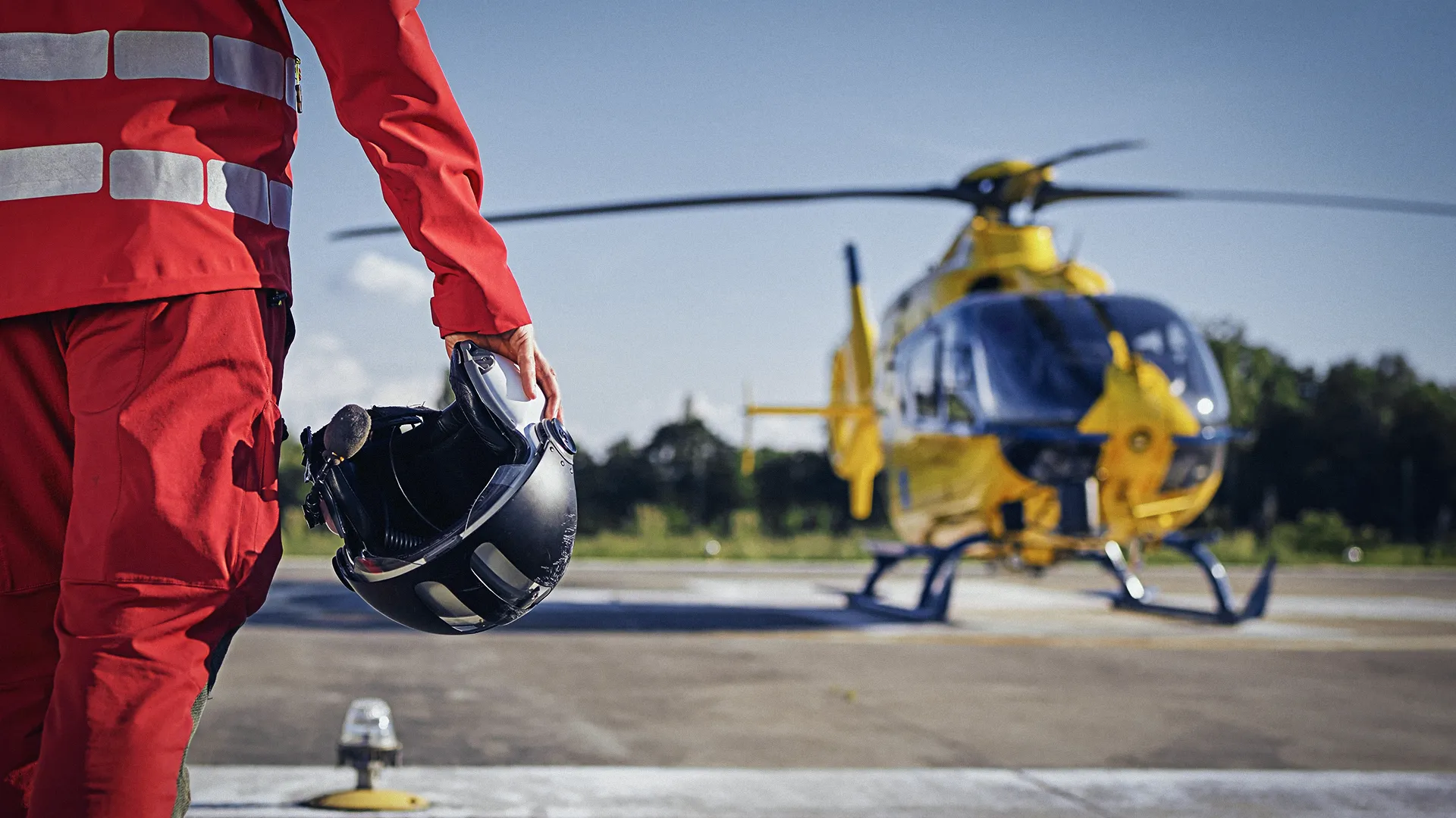 Eine Person in roter Schutzkleidung hält einen schwarzen Helm und steht auf einem Hubschrauberlandeplatz vor einem gelb-blauen Rettungshubschrauber.