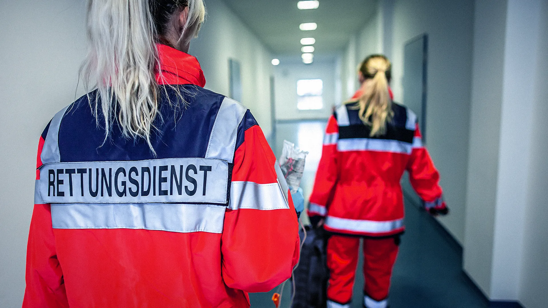 Zwei Personen in roten Rettungsdienst-Uniformen gehen einen Flur entlang, wobei eine Person im Vordergrund steht und die andere weiter vorne geht.
