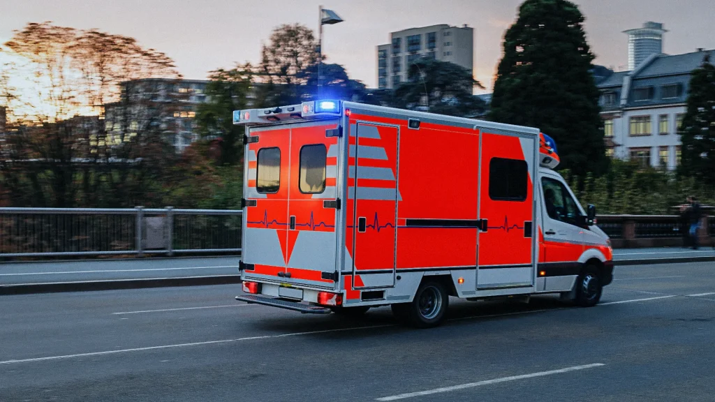 Ein leuchtend orangefarbenes Rettungsfahrzeug mit Blaulicht fährt auf einer Straße in einer städtischen Umgebung bei Dämmerung.