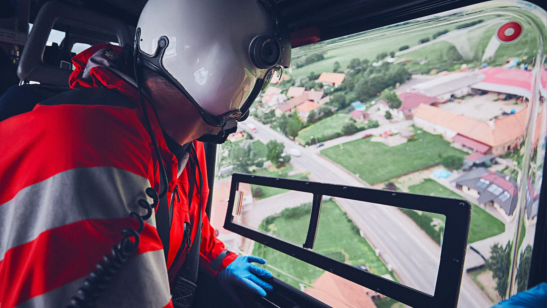 Eine Person in roter Schutzkleidung und Helm blickt aus dem Fenster eines Hubschraubers auf eine Landschaft mit Straßen und Gebäuden.