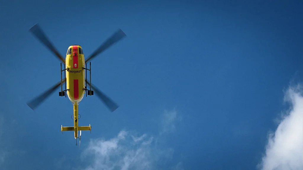 Ein gelb-roter Hubschrauber fliegt vor einem klaren blauen Himmel mit wenigen Wolken.