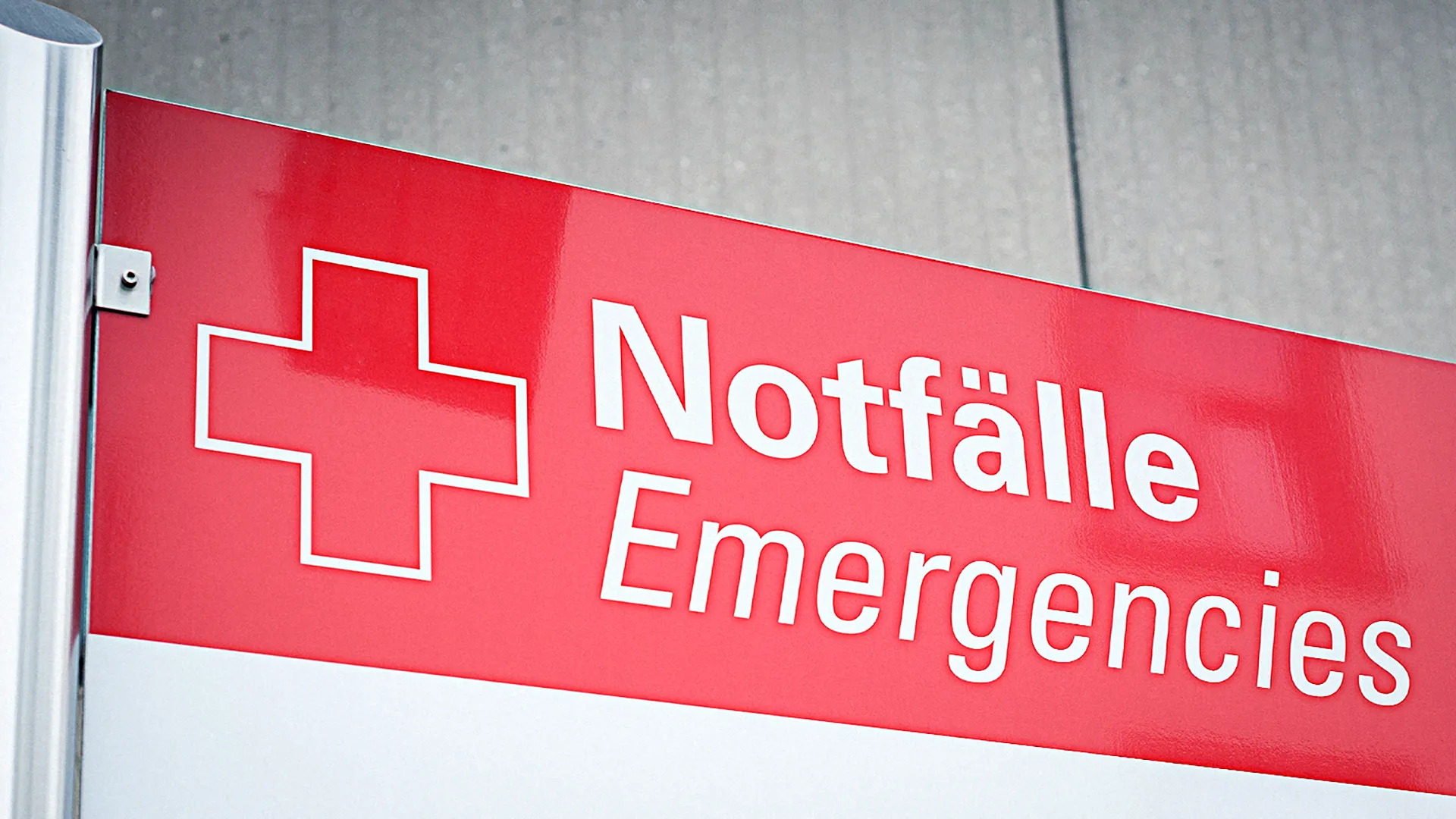 Ein rotes Schild mit einem weißen Kreuz und der Aufschrift Notfälle Emergencies an einer Gebäudewand.