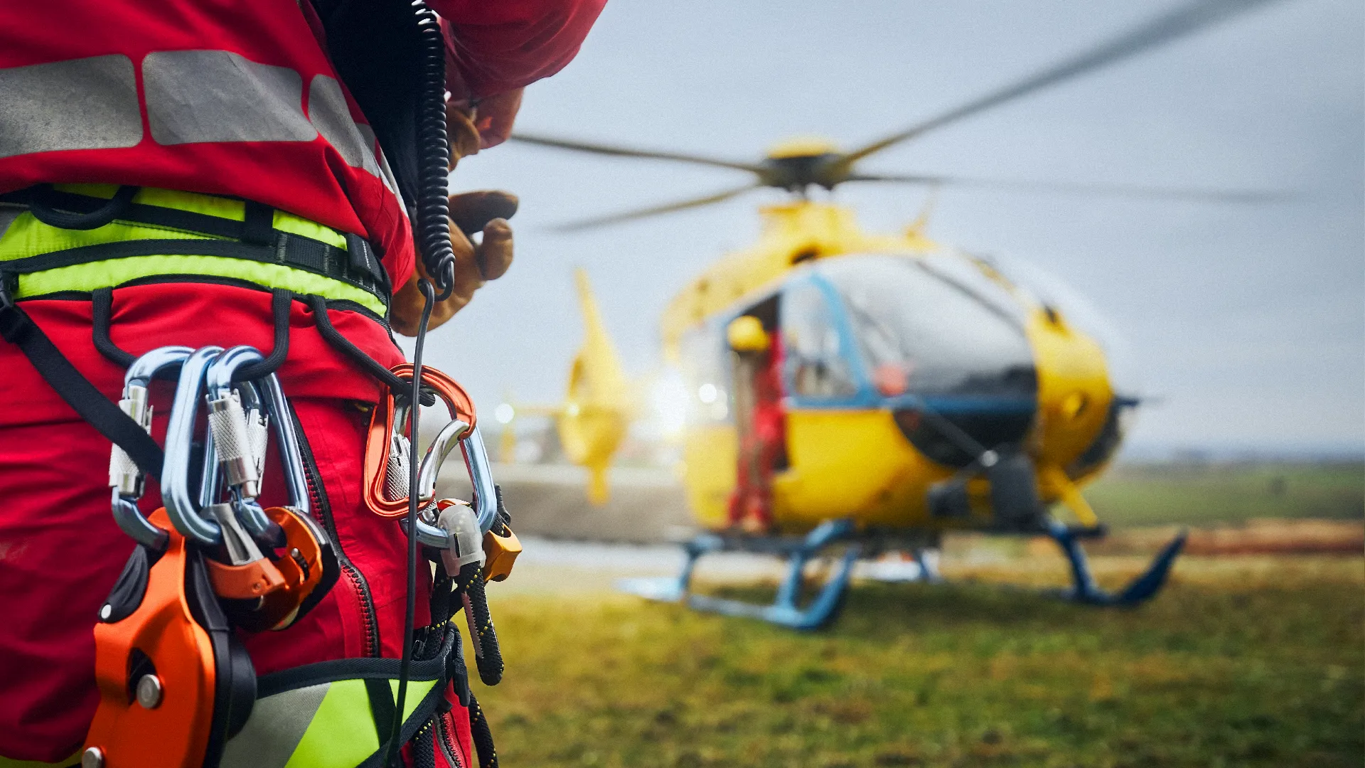 Eine Person in roter und gelber Schutzkleidung mit Kletterausrüstung steht auf einer Wiese vor einem gelben Rettungshubschrauber.