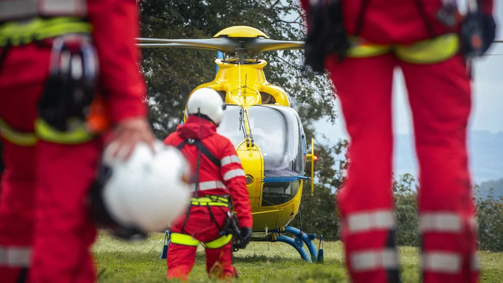 Mehrere Personen in roten Einsatzkleidung nähern sich einem gelben Rettungshubschrauber auf einer Wiese.